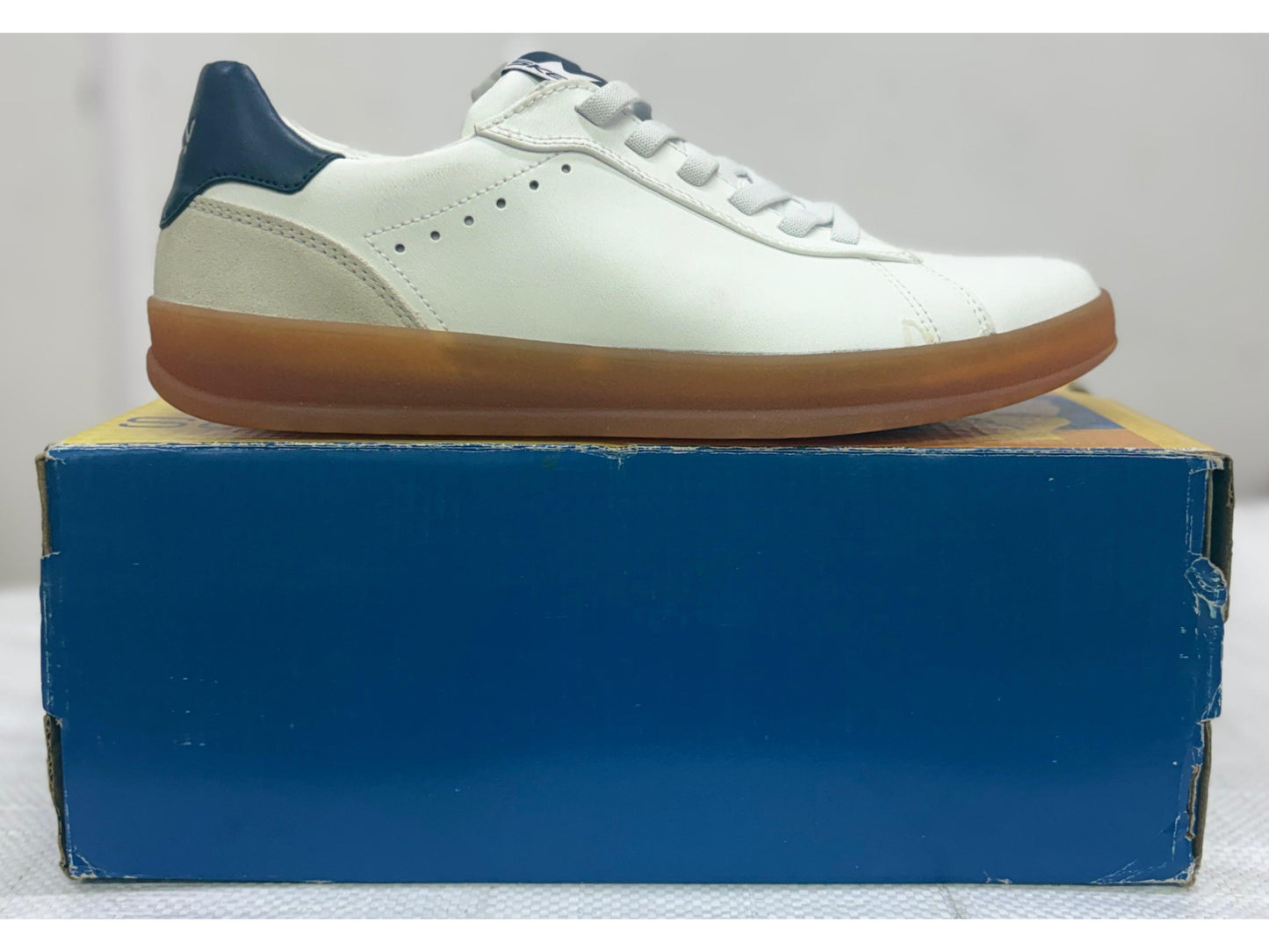 Side profile of a white Skechers leather sneaker with navy heel tab and caramel gum rubber sole on a blue box