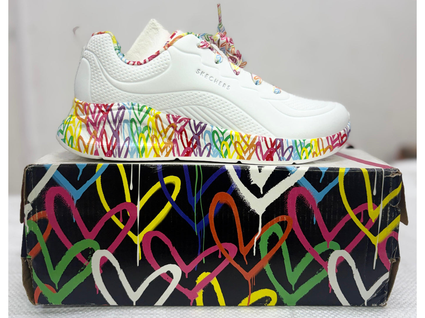 White Skechers lifestyle sneaker with multicolour graffiti heart print on midsole and laces on matching heart-pattern box.
