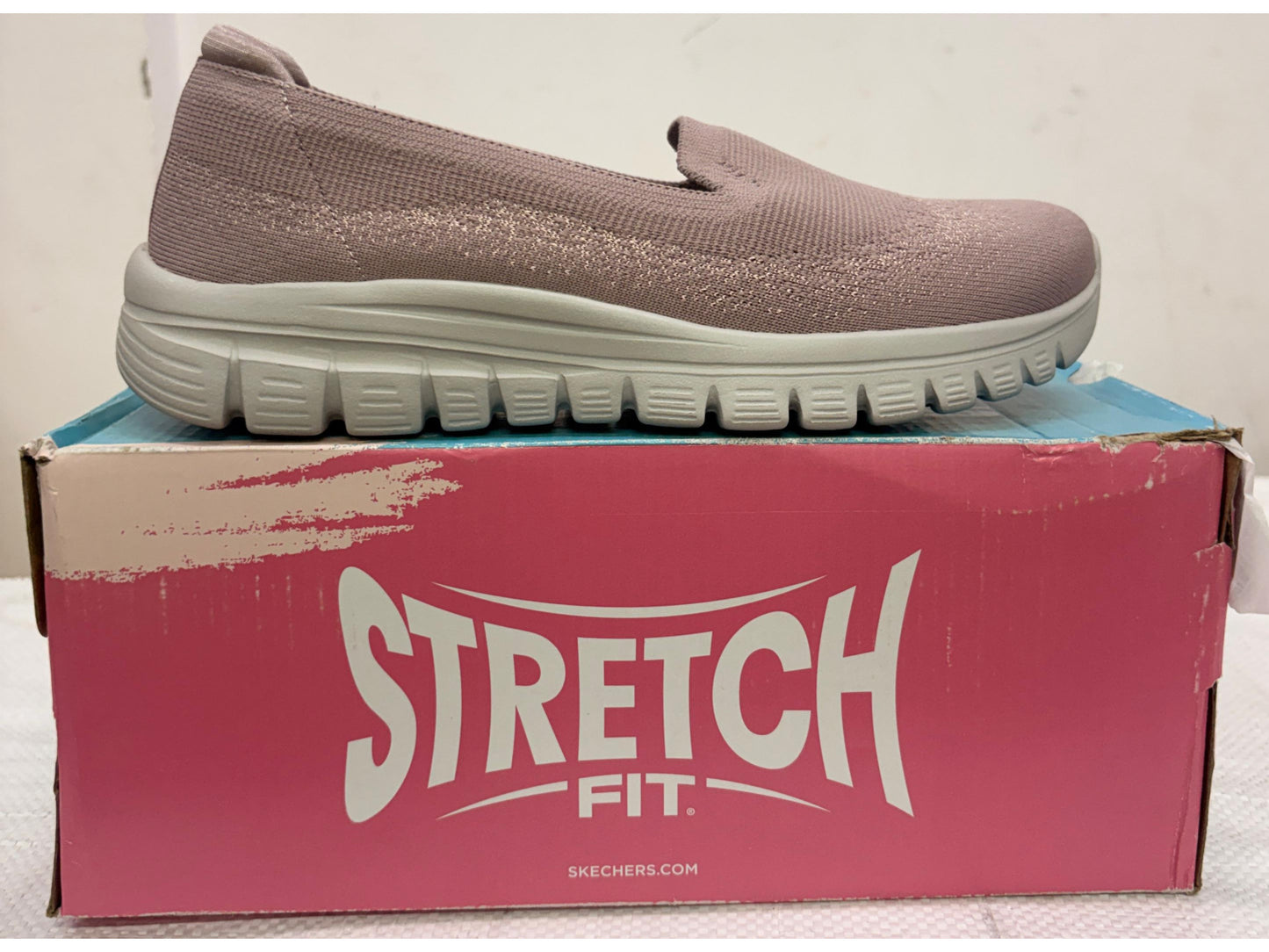 Side view of a mauve Skechers Stretch Fit slip-on walking shoe with light grey segmented sole on pink box